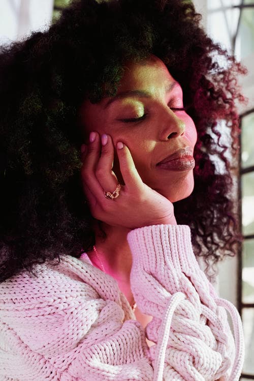 Emma Dabiri in white knit cardigan and white dress sat by window with subtle pink and green light from stained glass. She is wearing statement earrings, necklace and Claddagh ring.