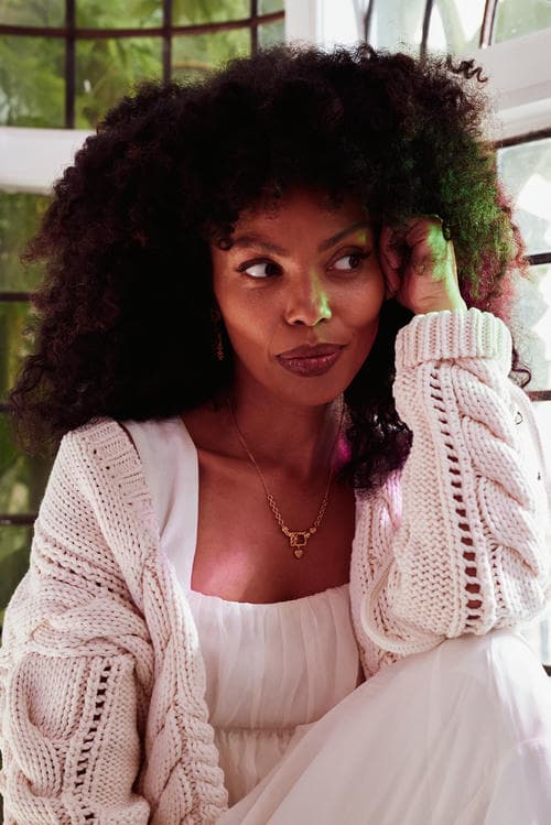 Emma Dabiri in white knit cardigan and white dress sat by window with subtle pink and green light from stained glass. She is wearing statement earrings, necklace and Claddagh ring.