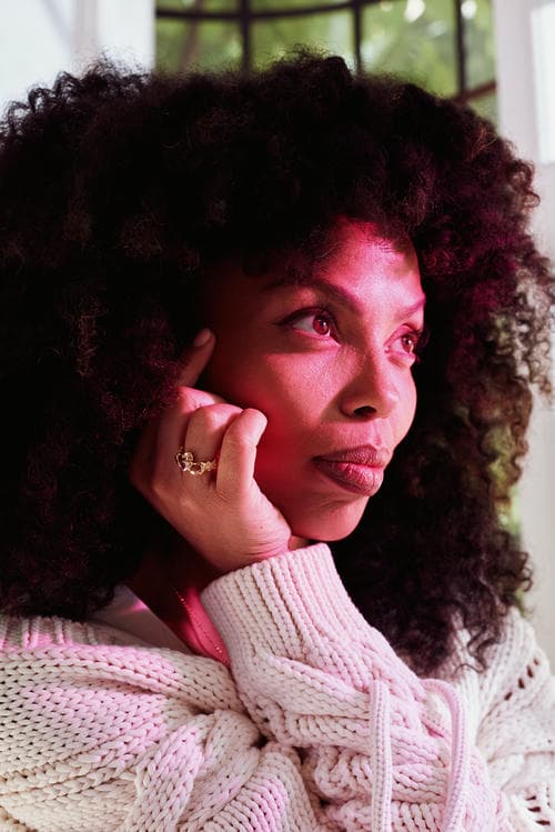 Emma Dabiri in white knit cardigan and white dress sat by window with subtle pink and green light from stained glass. She is wearing statement earrings, necklace and Claddagh ring.