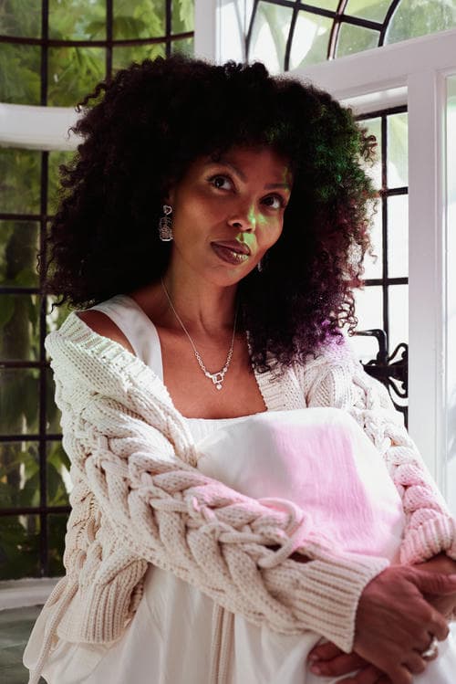 Emma Dabiri in white knit cardigan and white dress sat by window with subtle pink and green light from stained glass. She is wearing statement earrings, necklace and Claddagh ring.
